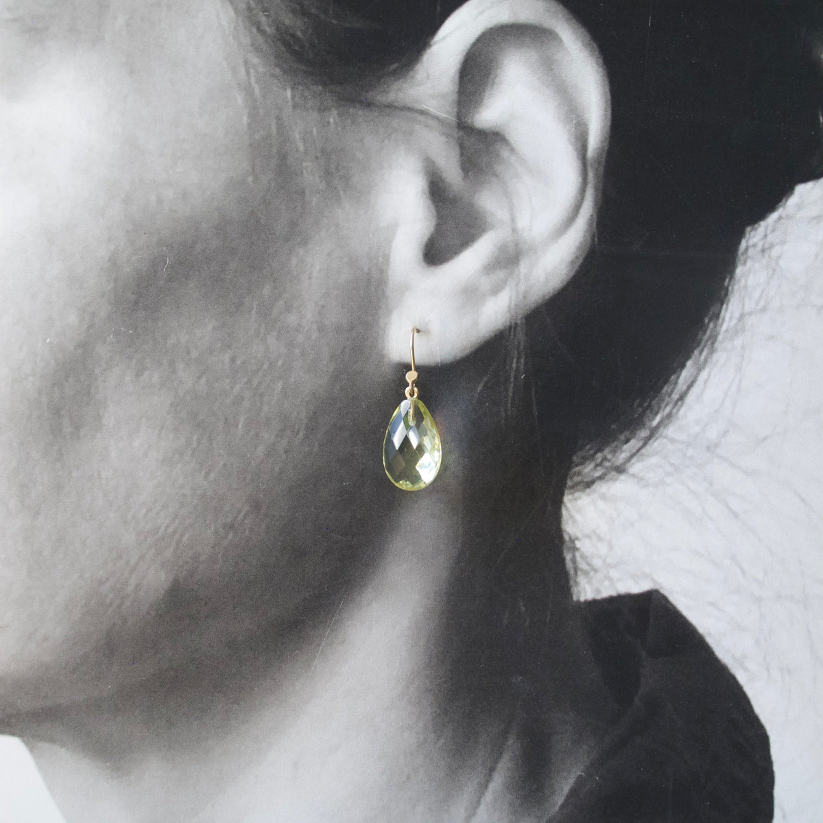 A close-up of a person's ear wearing a small gold teardrop earring with a faceted yellow-green stone.