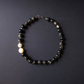 A beaded bracelet made of mostly dark black and smoky gray round beads, with two smooth off-white beads on one side and a silver clasp at the top, photographed on a dark gray background.
