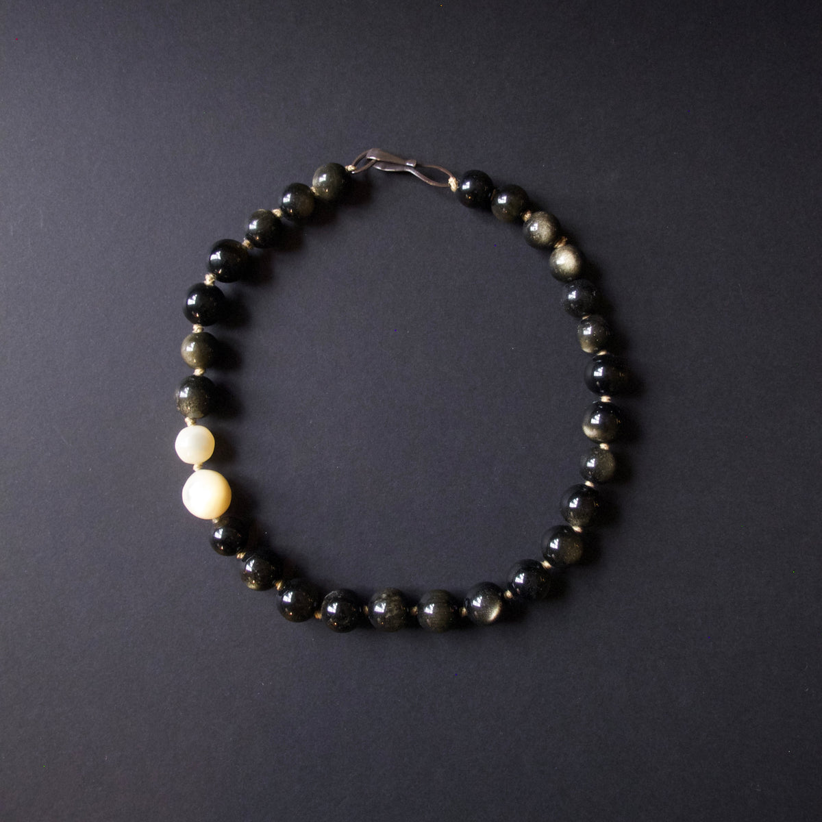 A beaded bracelet made of mostly dark black and smoky gray round beads, with two smooth off-white beads on one side and a silver clasp at the top, photographed on a dark gray background.