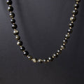 A beaded necklace made of polished dark gray to black round beads with subtle metallic gold flecks, displayed against a dark gray background.