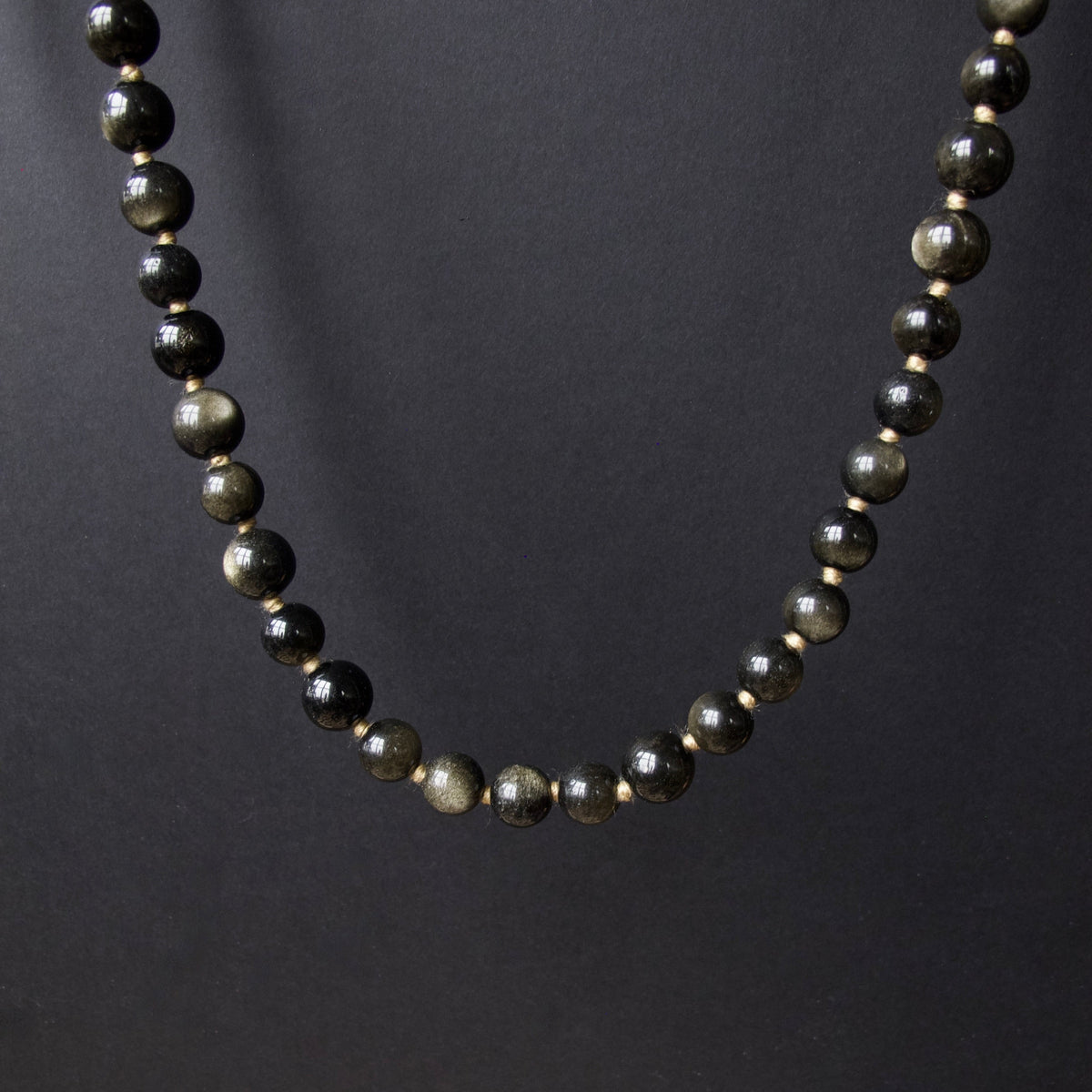 A beaded necklace made of polished dark gray to black round beads with subtle metallic gold flecks, displayed against a dark gray background.