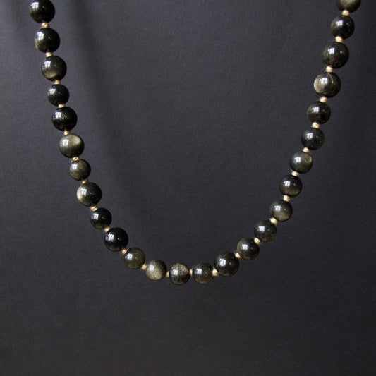 A beaded necklace made of polished dark gray to black round beads with subtle metallic gold flecks, displayed against a dark gray background.