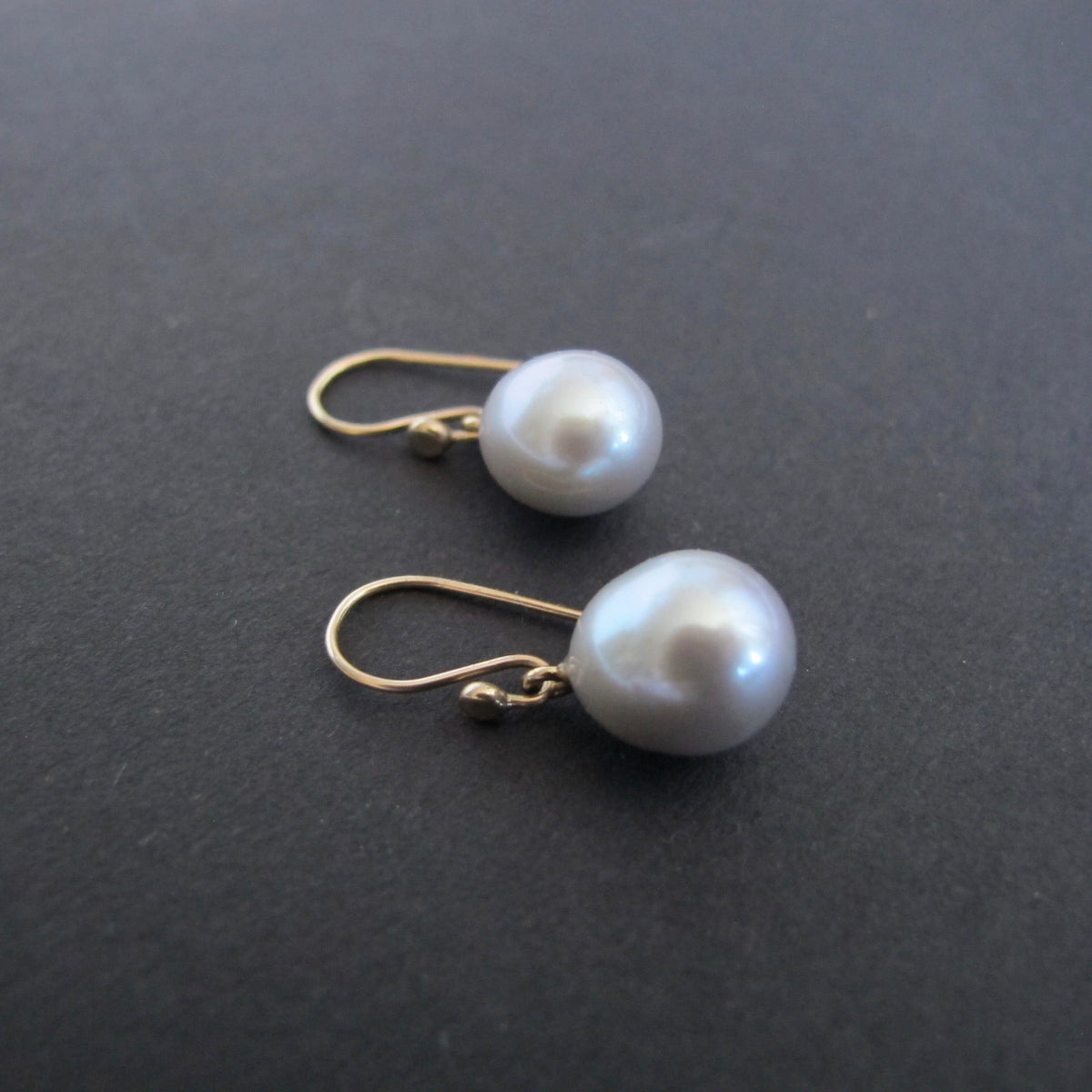 A pair of drop earrings featuring lustrous white freshwater pearls suspended from gold French hook wires, displayed on a dark gray textured surface.
