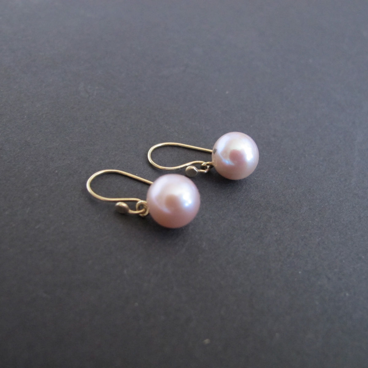 A pair of gold fishhook drop earrings with smooth, lustrous light pink freshwater pearls, displayed on a dark gray textured surface.