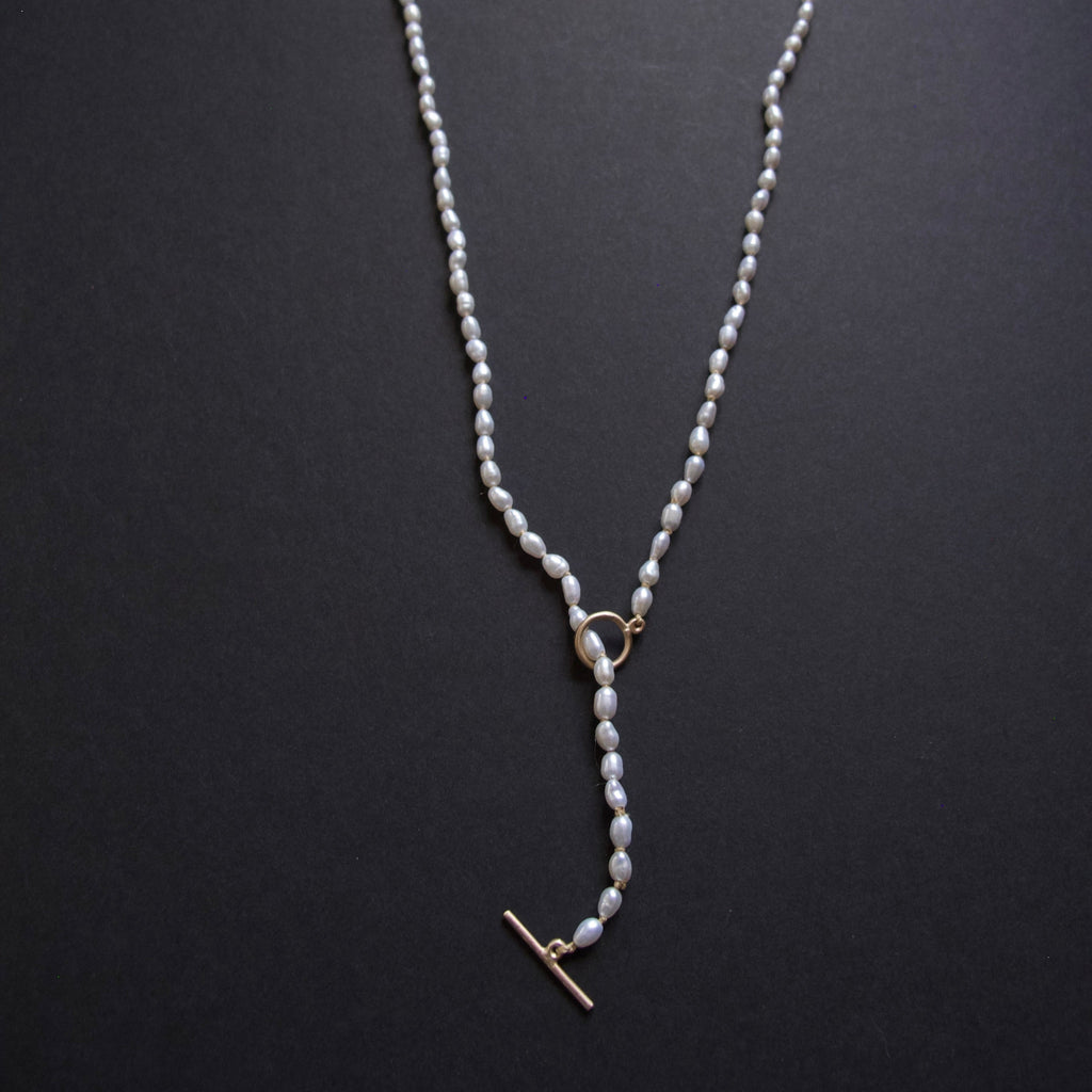 A delicate pearl necklace with a gold toggle clasp and ring connector, displayed on a dark textured background.