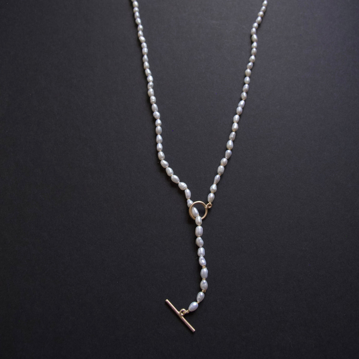 A delicate pearl necklace with a gold toggle clasp and ring connector, displayed on a dark textured background.