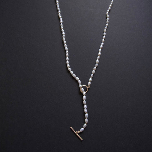 A delicate pearl necklace with a gold toggle clasp and ring connector, displayed on a dark textured background.