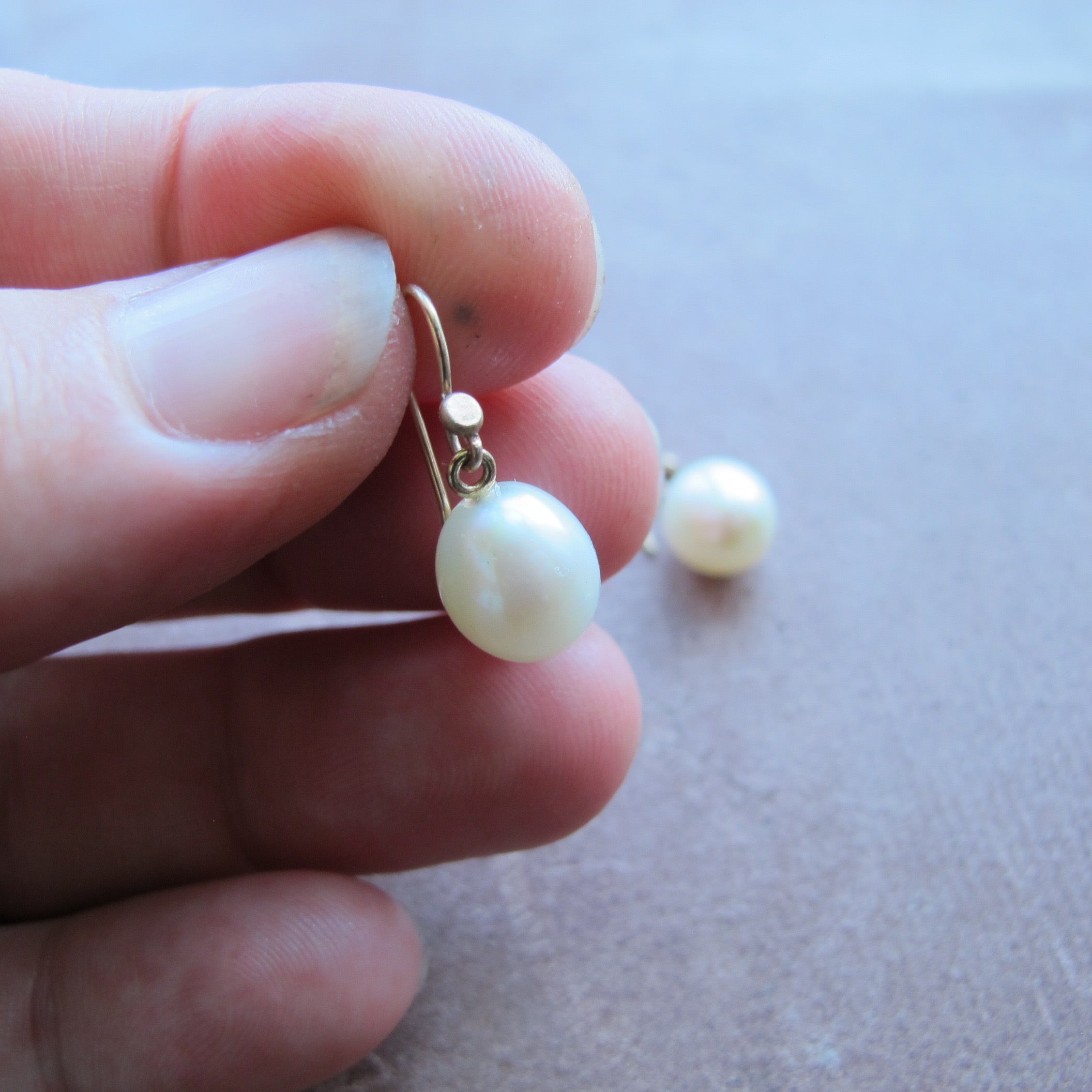 A close-up of a hand holding a small gold hoop earring with a single round white pearl, with a second matching earring resting on a neutral surface in the background.