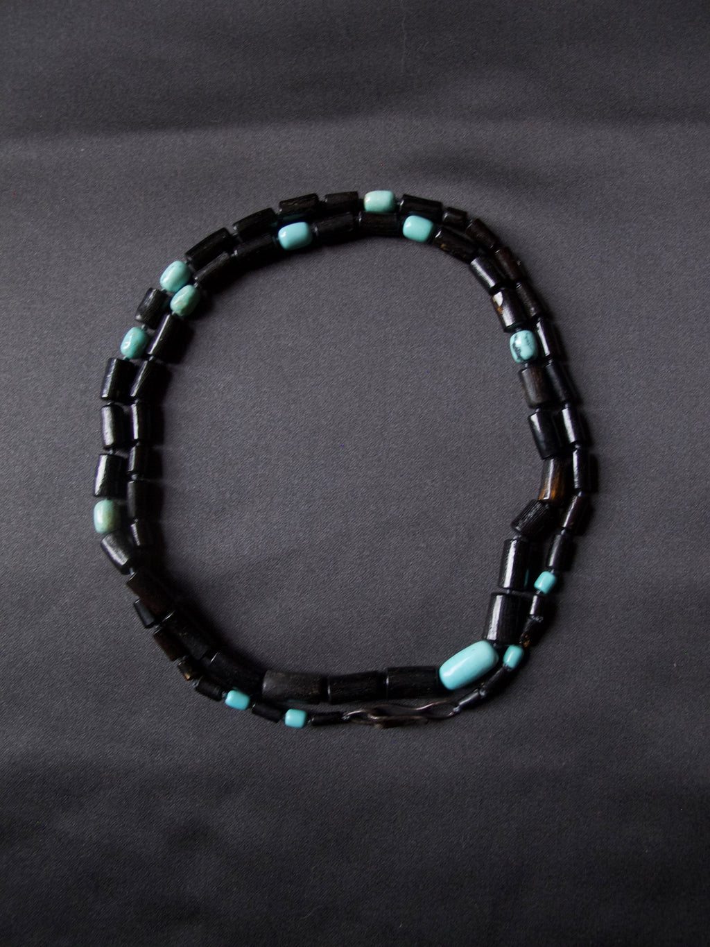 A beaded necklace made of dark black cylindrical beads with small turquoise blue spacer beads, arranged in an oval shape on a dark gray background.