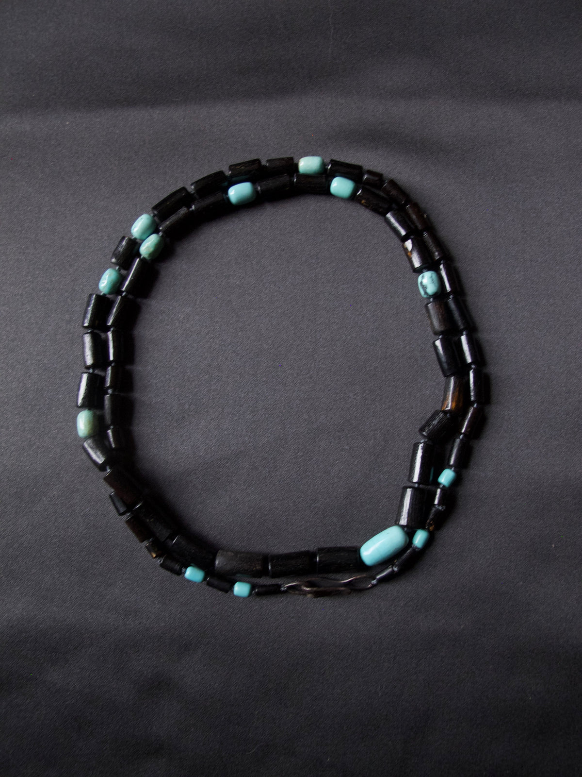 A beaded necklace made of dark black cylindrical beads with small turquoise blue spacer beads, arranged in an oval shape on a dark gray background.
