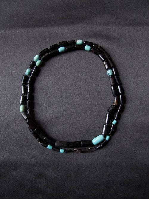 A beaded necklace made of dark black cylindrical beads with small turquoise blue spacer beads, arranged in an oval shape on a dark gray background.