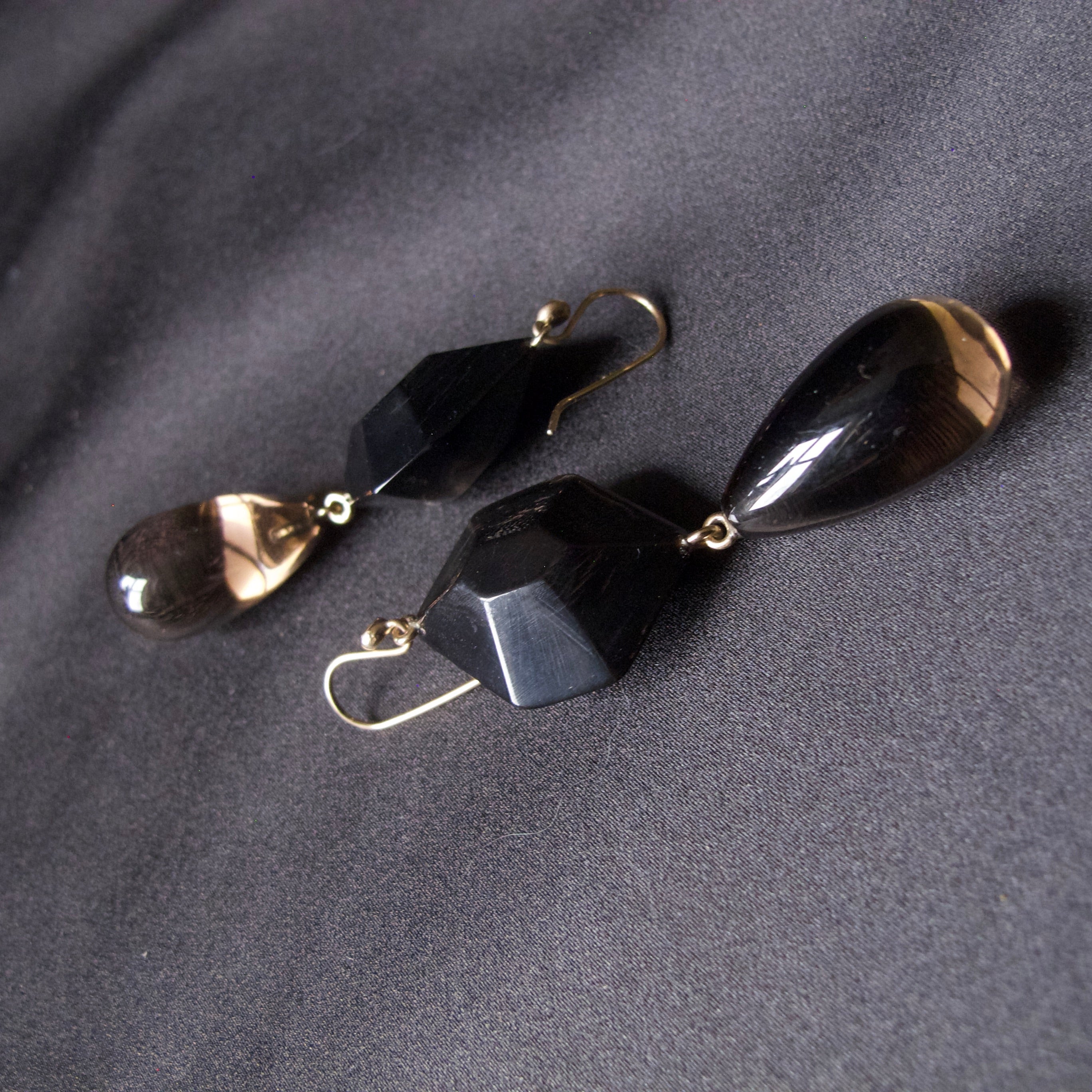 A pair of drop earrings with faceted black beads and teardrop-shaped smoky brown glass drops, attached to gold hooks, displayed on a dark gray fabric background.