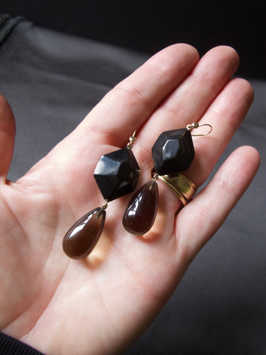 A pair of handmade drop earrings held in a person's hand. The earrings feature faceted black gemstone beads at the top and teardrop-shaped dark brown glass drops at the bottom, suspended from gold-toned wires.