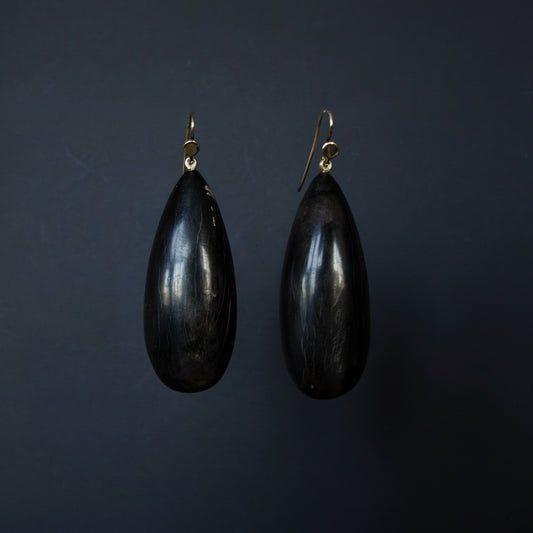 A pair of teardrop-shaped black wooden earrings with gold fishhook wires, displayed against a dark gray background.
