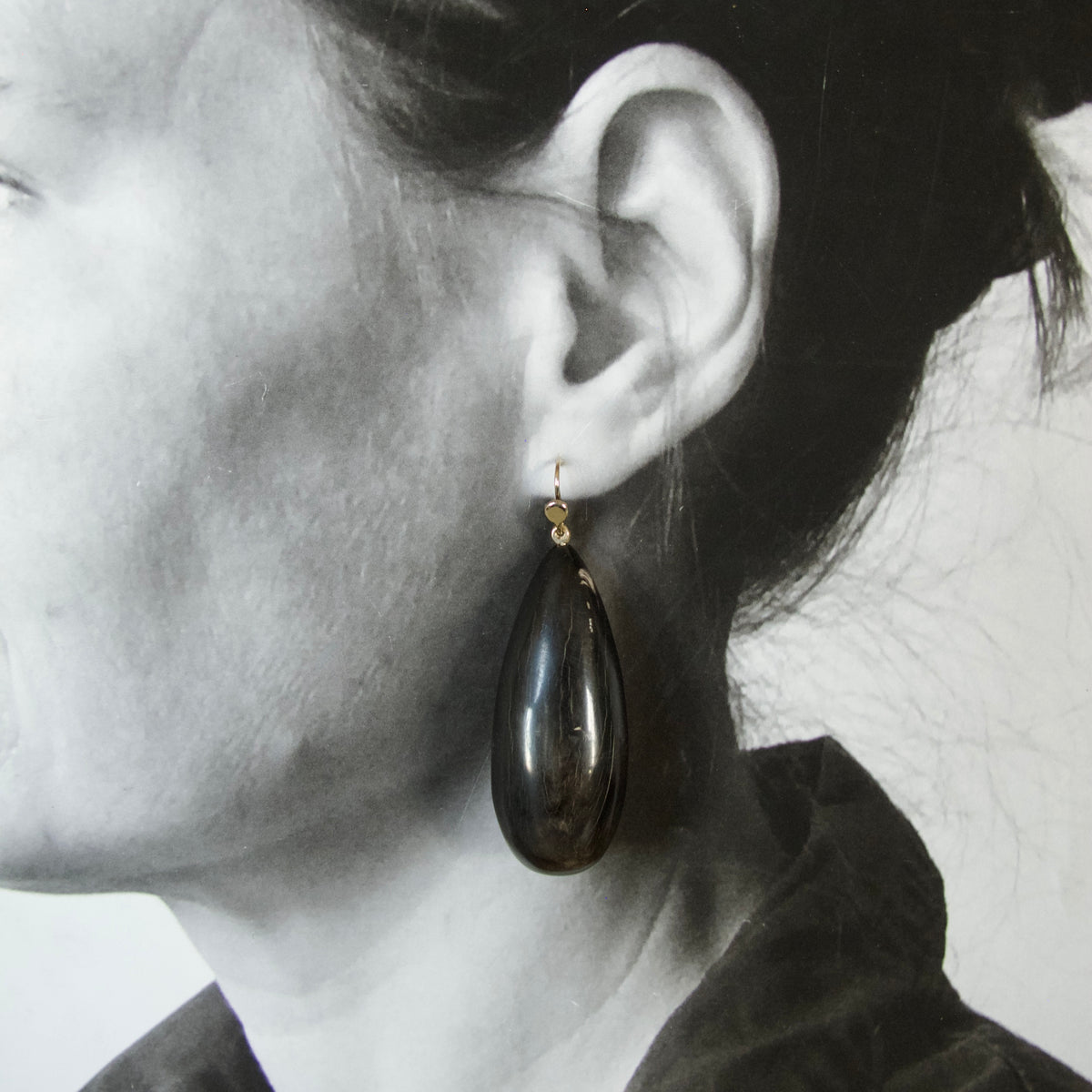 A close-up side profile of a woman wearing a dark teardrop-shaped dangle earring with a small gold hook.