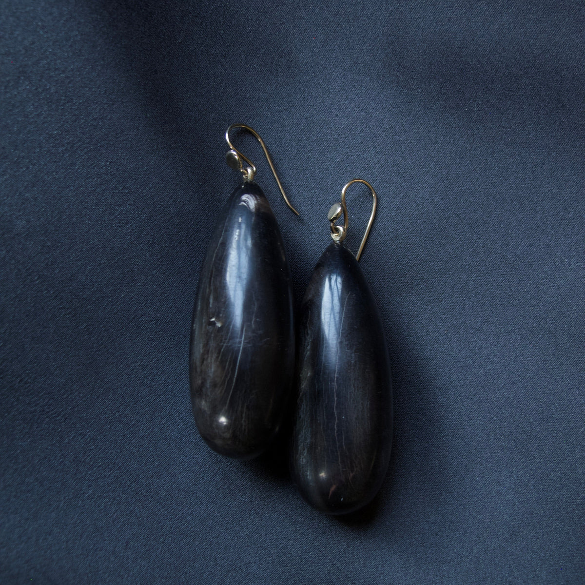 A pair of dark teardrop-shaped earrings with gold-tone fishhook wires, displayed on a dark navy blue fabric background.