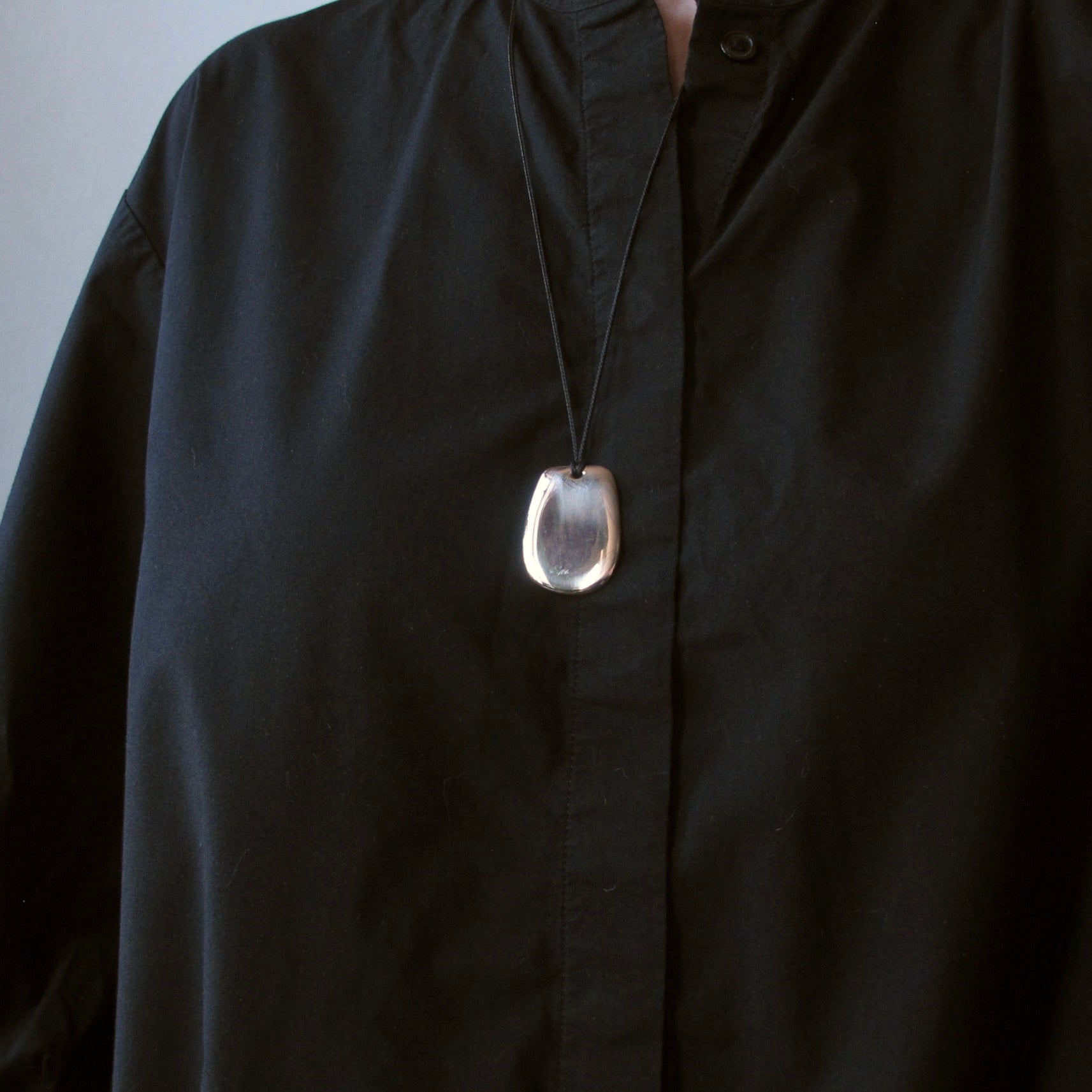 A close-up of a person wearing a black button-up shirt and a black cord necklace with a polished, teardrop-shaped silver pendant.