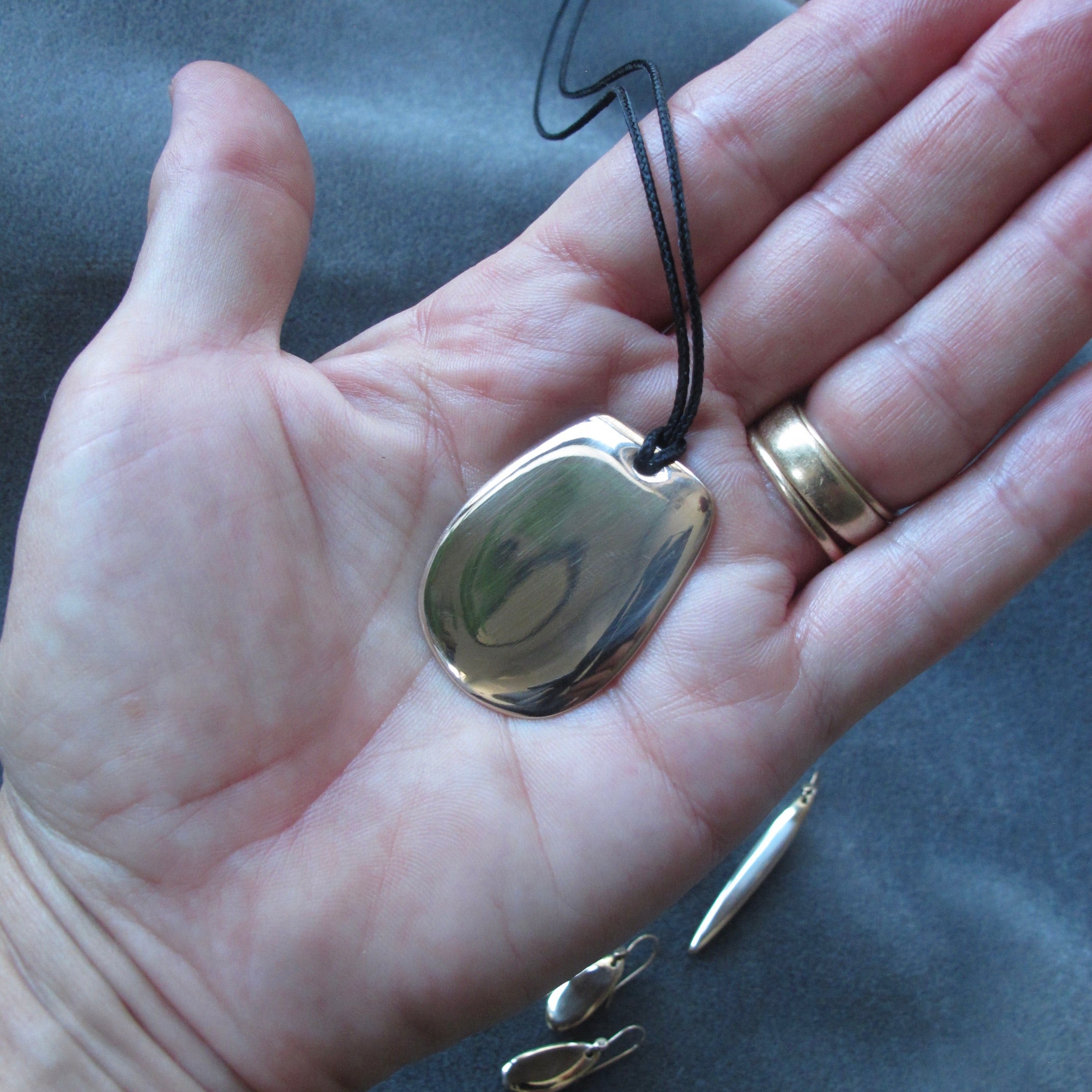 A polished gold-toned pendant necklace with an irregular organic shape, hanging from a black cord and displayed on an open hand. A gold ring is visible on the model's finger, and other jewelry pieces are partially visible at the bottom against a dark gray fabric background.