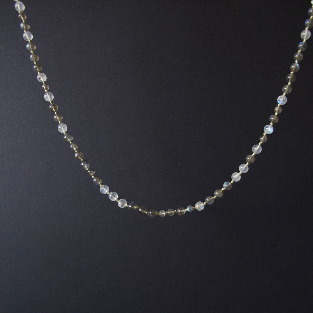 A delicate beaded necklace with small iridescent white beads and tiny gold spacer beads, displayed against a dark black background.