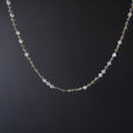 A delicate beaded necklace with small iridescent white beads and tiny gold spacer beads, displayed against a dark black background.