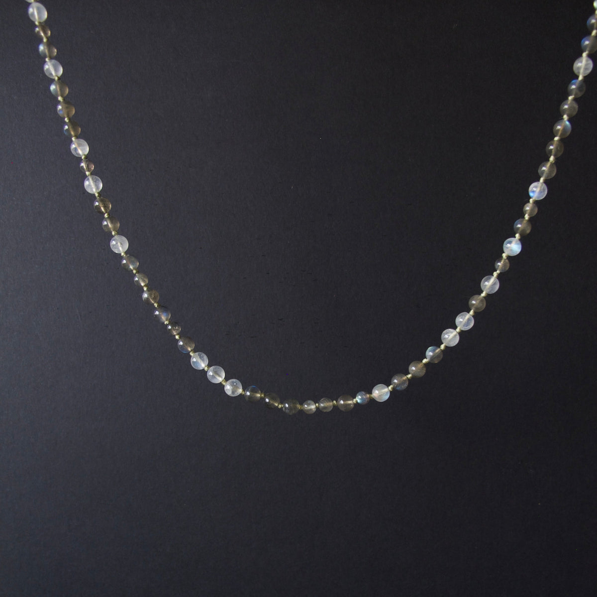 A delicate beaded necklace with small iridescent white beads and tiny gold spacer beads, displayed against a dark black background.