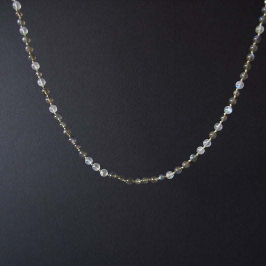 A delicate beaded necklace with small iridescent white beads and tiny gold spacer beads, displayed against a dark black background.