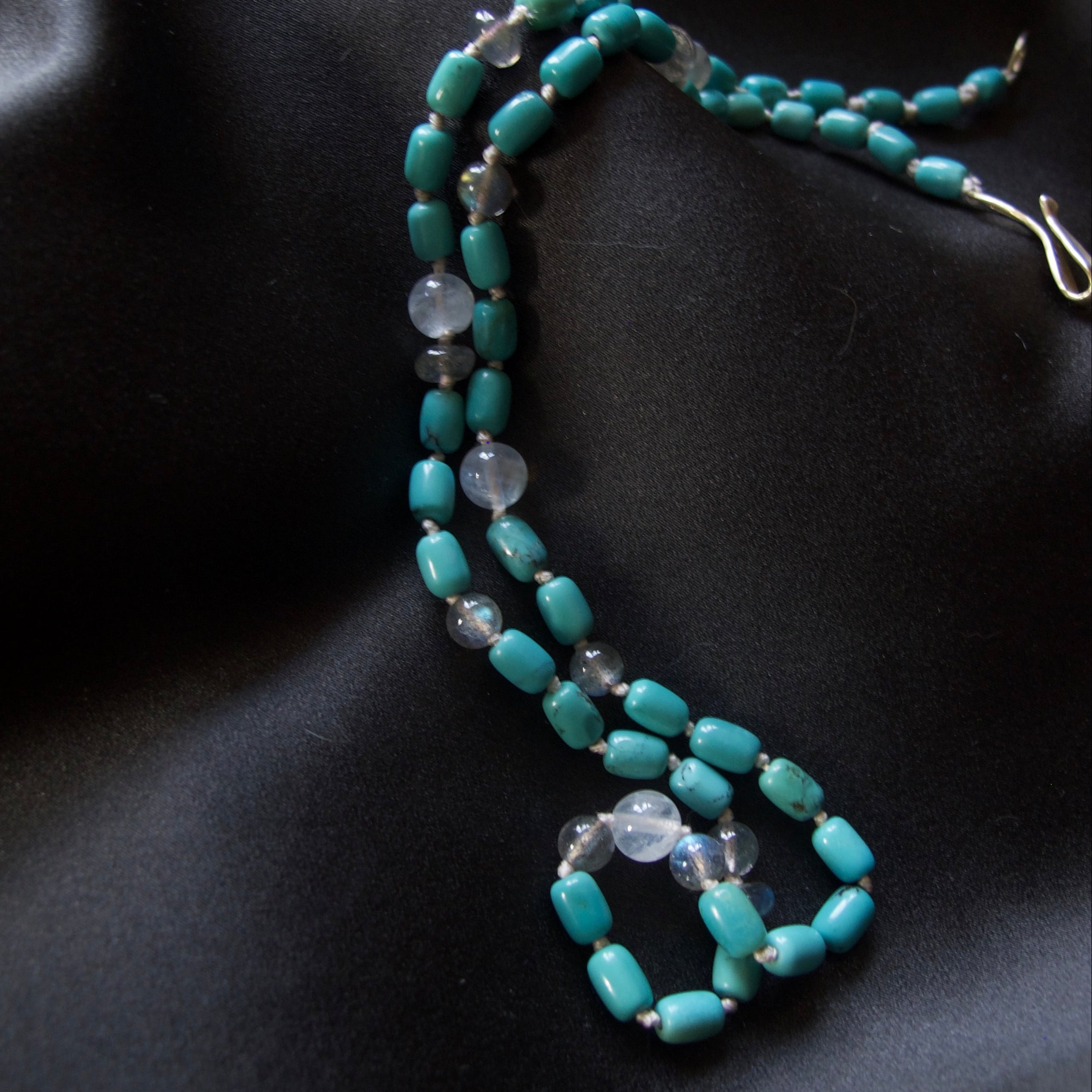 A beaded necklace with turquoise-blue cylindrical beads and translucent white oval beads, displayed on a draped black satin fabric.