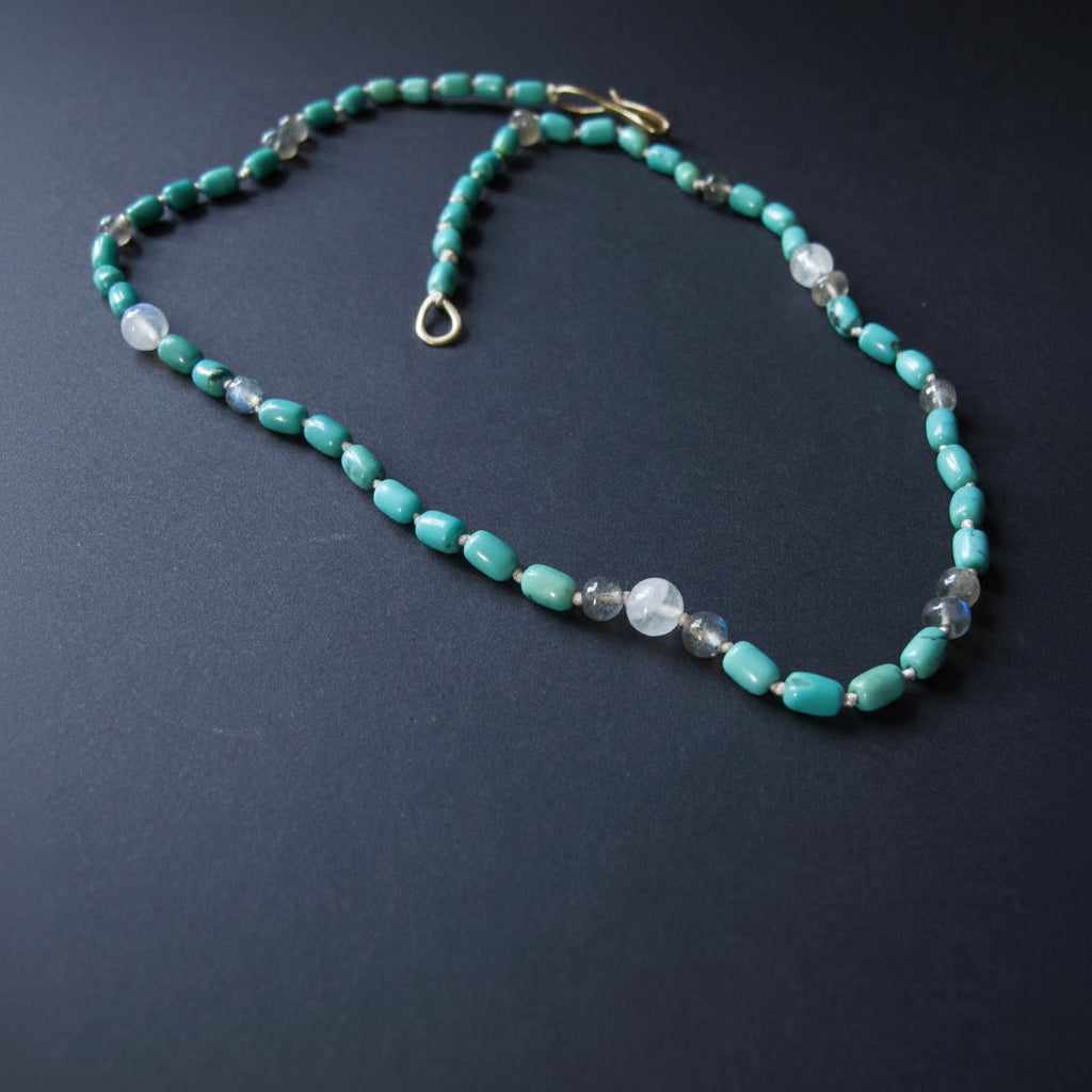A beaded necklace featuring turquoise-colored rectangular beads, clear faceted round beads, and small dark gray beads, with a gold clasp, displayed on a dark gray background.