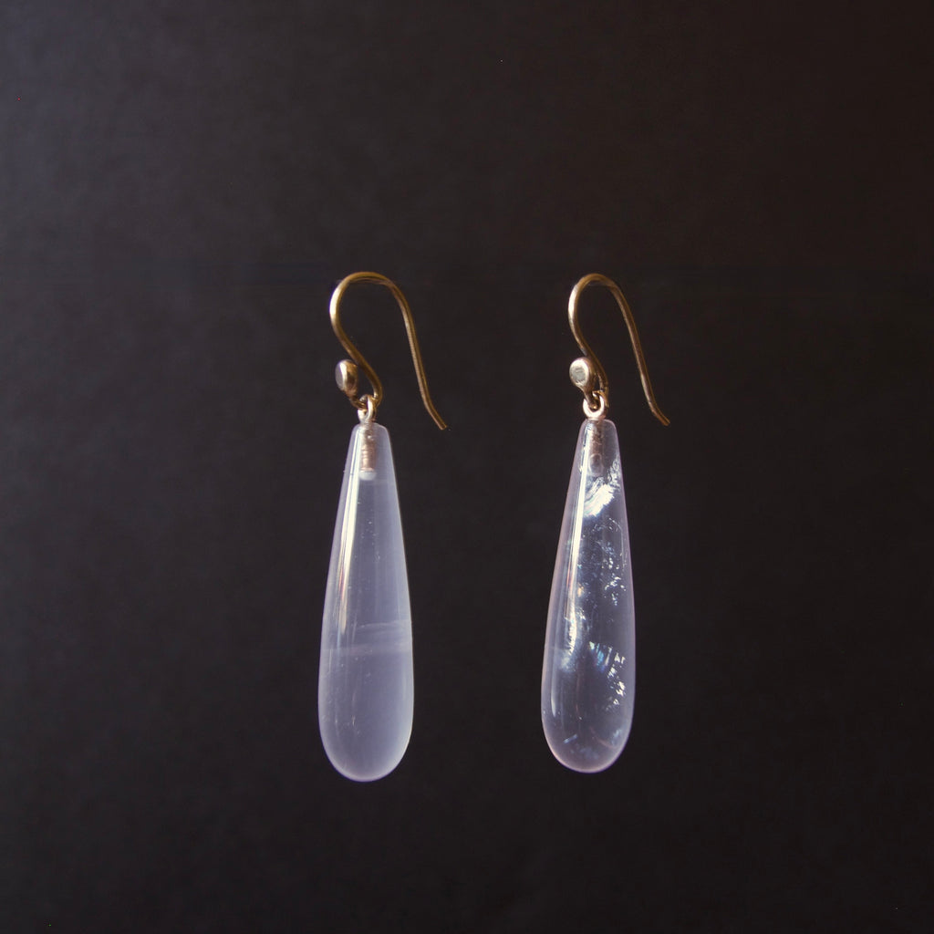 A pair of teardrop-shaped drop earrings with milky translucent pale lavender-blue stones and gold hooks, shown against a dark black background.