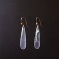 A pair of teardrop-shaped drop earrings with milky translucent pale lavender-blue stones and gold hooks, shown against a dark black background.