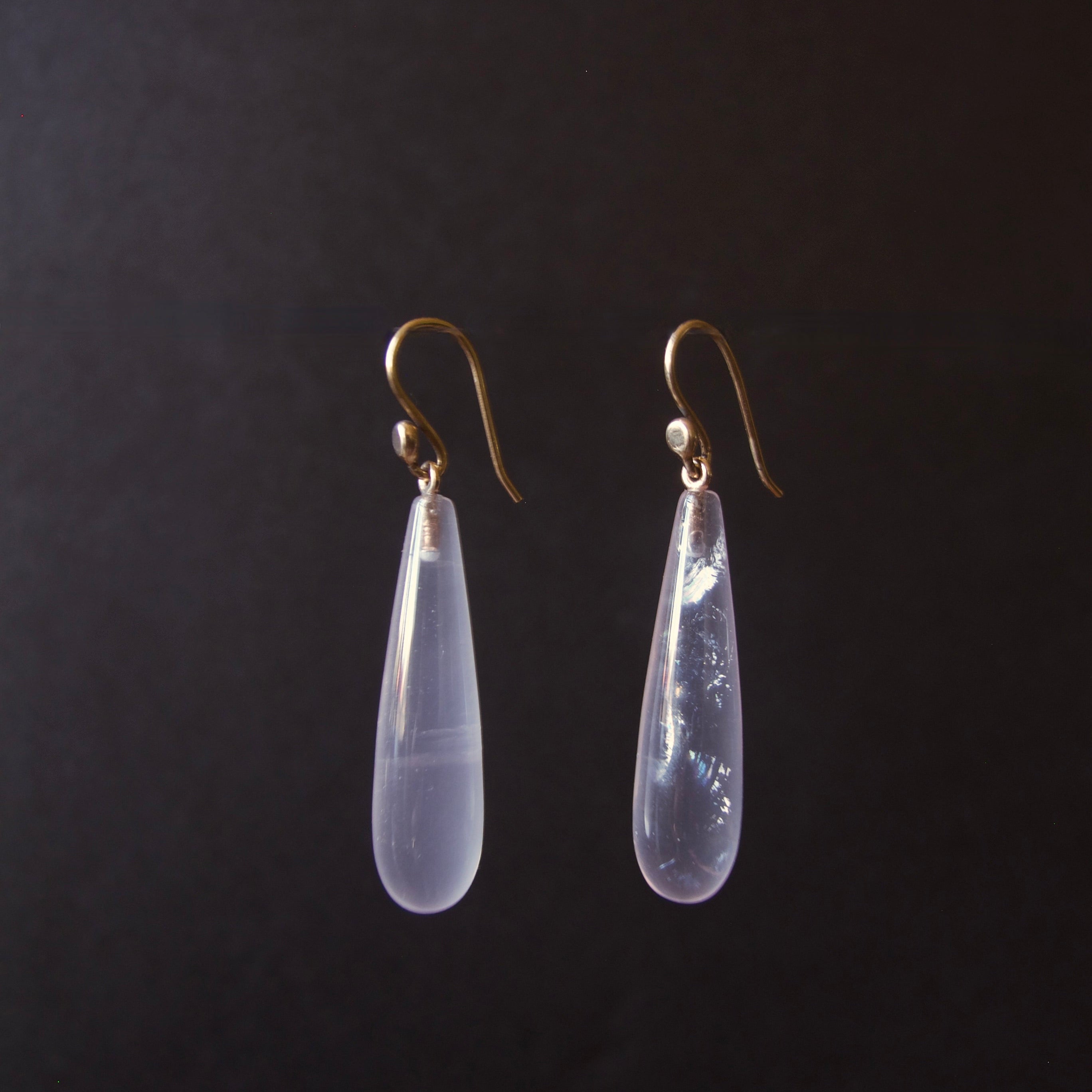 A pair of teardrop-shaped drop earrings with milky translucent pale lavender-blue stones and gold hooks, shown against a dark black background.
