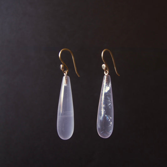 A pair of teardrop-shaped drop earrings with milky translucent pale lavender-blue stones and gold hooks, shown against a dark black background.