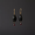 A pair of dangle earrings with glossy black teardrop-shaped beads and small dangling red teardrop gemstones, hanging from gold-tone hooks on a white display bar against a white background.
