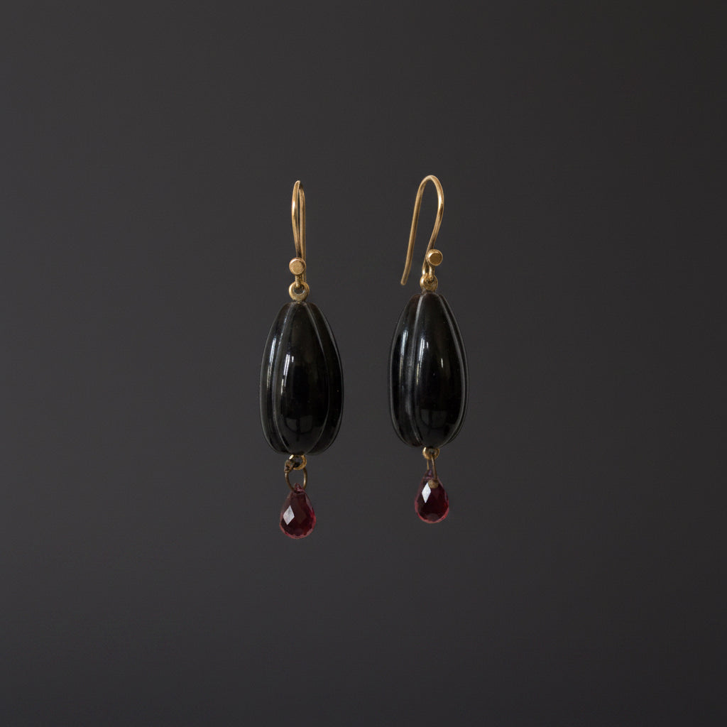 A pair of dangle earrings with glossy black teardrop-shaped beads and small dangling red teardrop gemstones, hanging from gold-tone hooks on a white display bar against a white background.