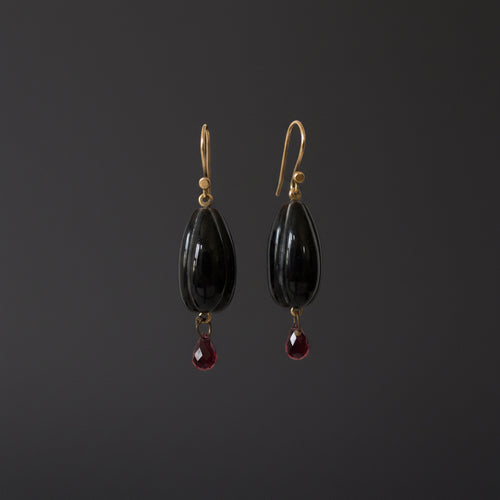 A pair of dangle earrings with glossy black teardrop-shaped beads and small dangling red teardrop gemstones, hanging from gold-tone hooks on a white display bar against a white background.