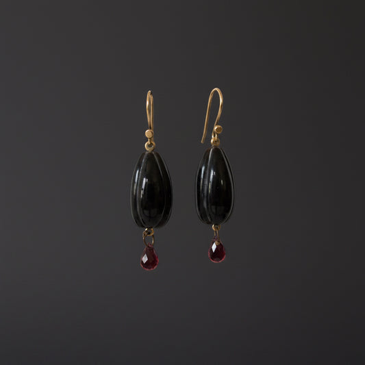 A pair of dangle earrings with glossy black teardrop-shaped beads and small dangling red teardrop gemstones, hanging from gold-tone hooks on a white display bar against a white background.