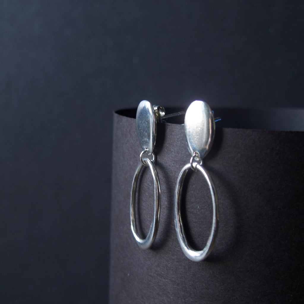 A pair of silver drop earrings with oval dangles, displayed on a black cylindrical stand against a dark background.