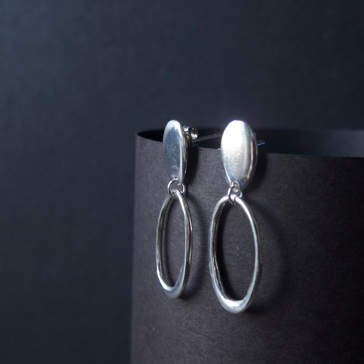A pair of silver drop earrings with oval dangles, displayed on a black cylindrical stand against a dark background.