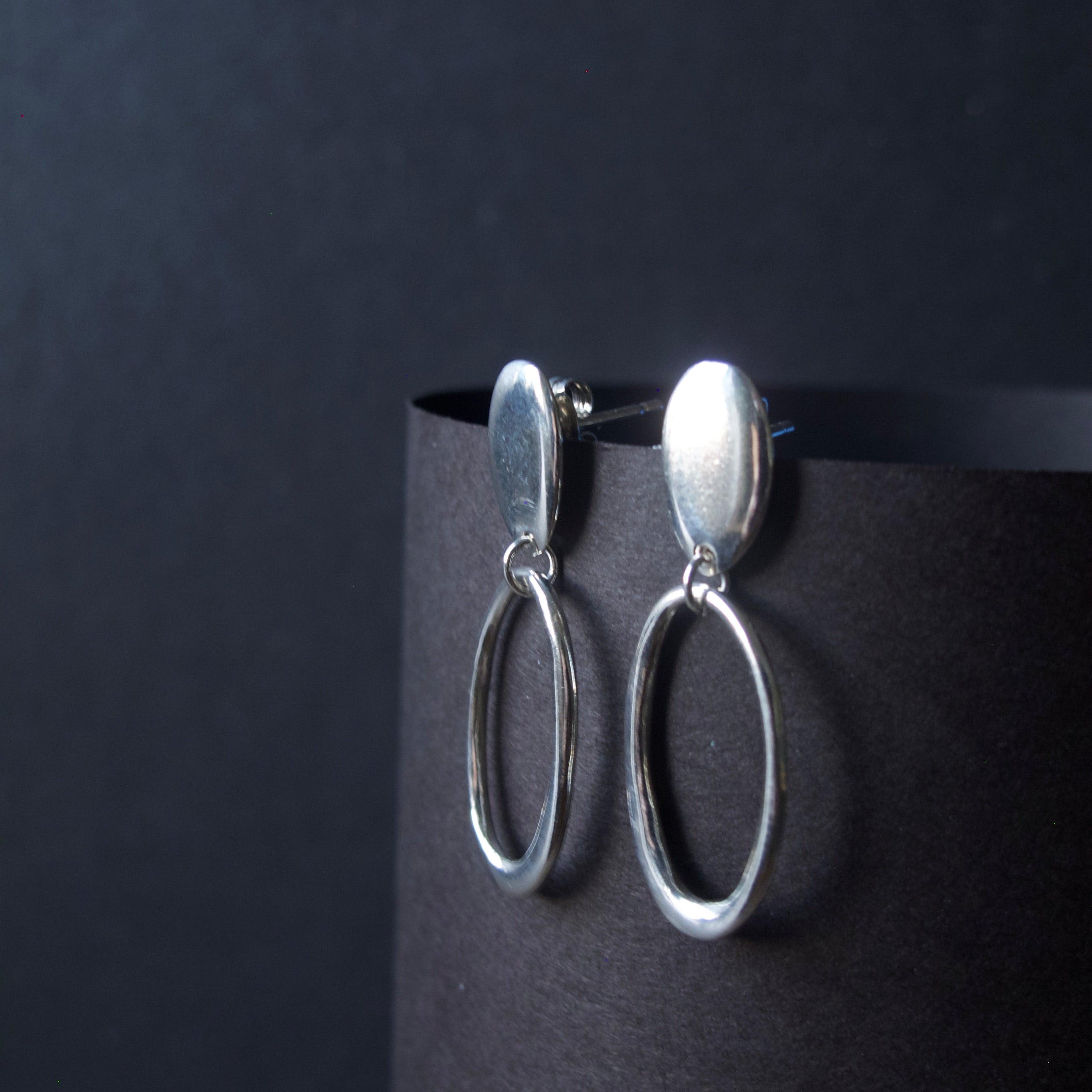 A pair of silver drop earrings with oval dangles, displayed on a black cylindrical stand against a dark background.