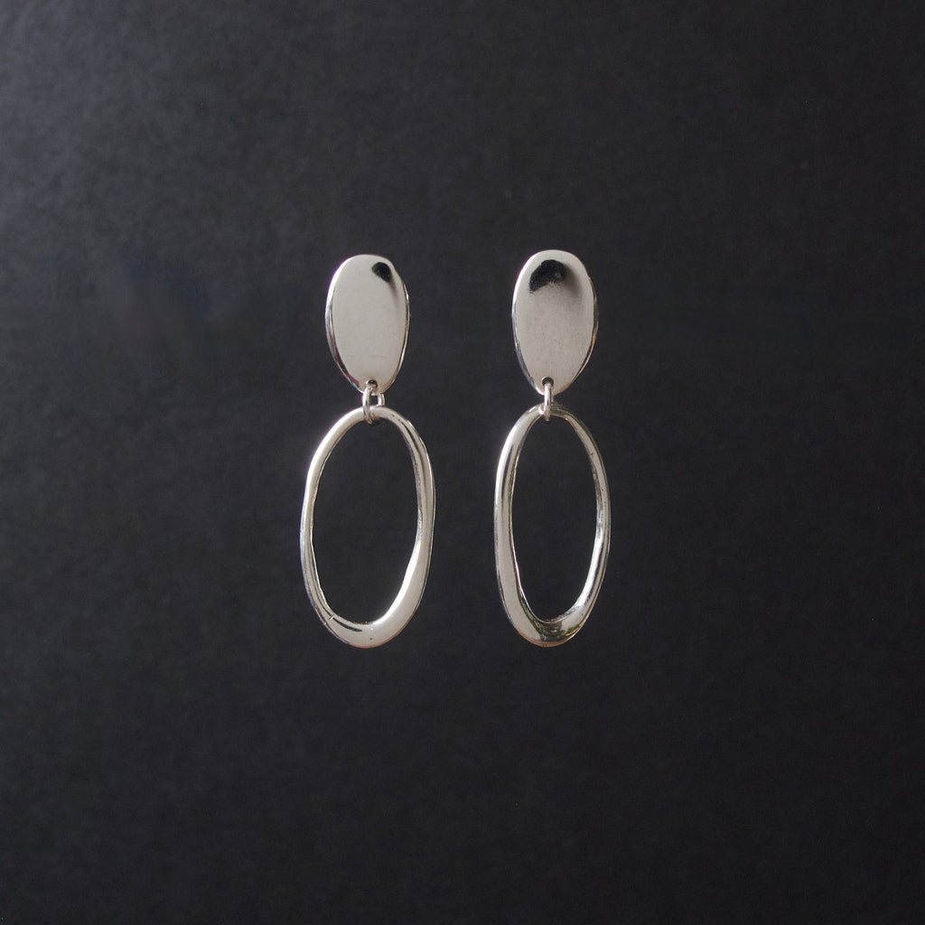A pair of silver drop earrings with a flat oval stud and a larger textured oval dangle, displayed on a dark textured background.