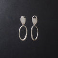 A pair of silver drop earrings with a flat oval stud and a larger textured oval dangle, displayed on a dark textured background.