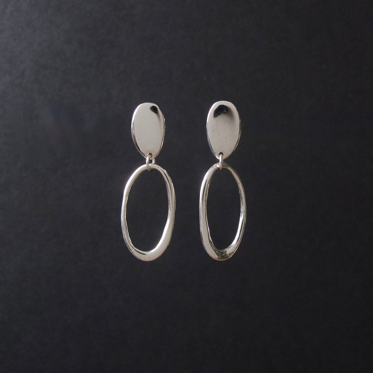 A pair of silver drop earrings with a flat oval stud and a larger textured oval dangle, displayed on a dark textured background.
