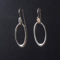 A pair of silver drop earrings with large open oval hoops and small flat circular accents, shown against a dark black background.