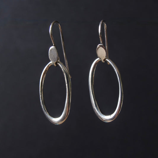 A pair of silver drop earrings with large open oval hoops and small flat circular accents, shown against a dark black background.