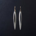 A pair of long, slender silver drop earrings with a polished finish and gold-tone hooks, hanging against a dark black background.