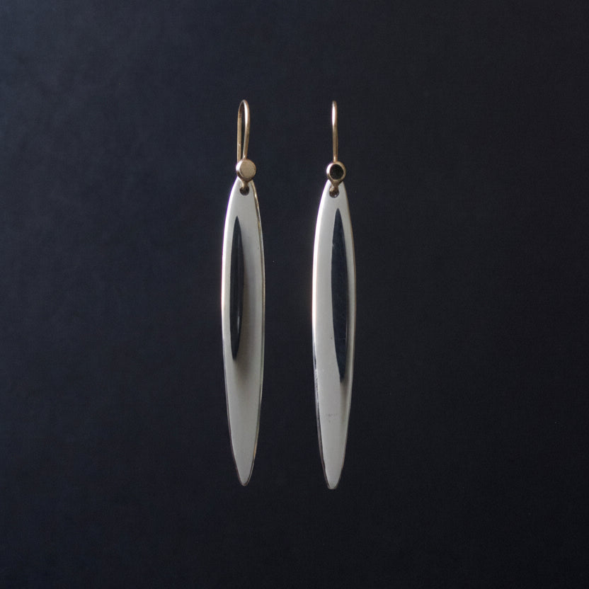A pair of long, slender silver drop earrings with a polished finish and gold-tone hooks, hanging against a dark black background.