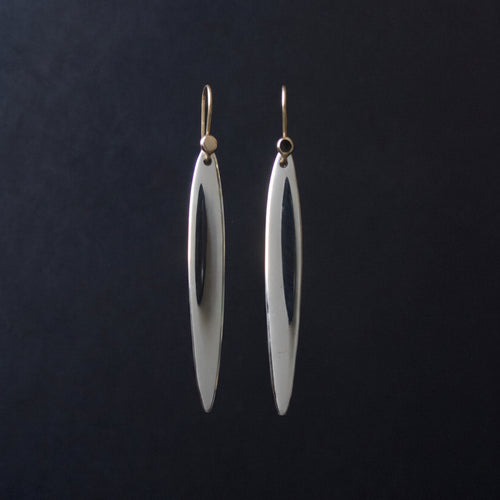 A pair of long, slender silver drop earrings with a polished finish and gold-tone hooks, hanging against a dark black background.