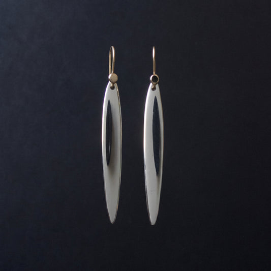 A pair of long, slender silver drop earrings with a polished finish and gold-tone hooks, hanging against a dark black background.
