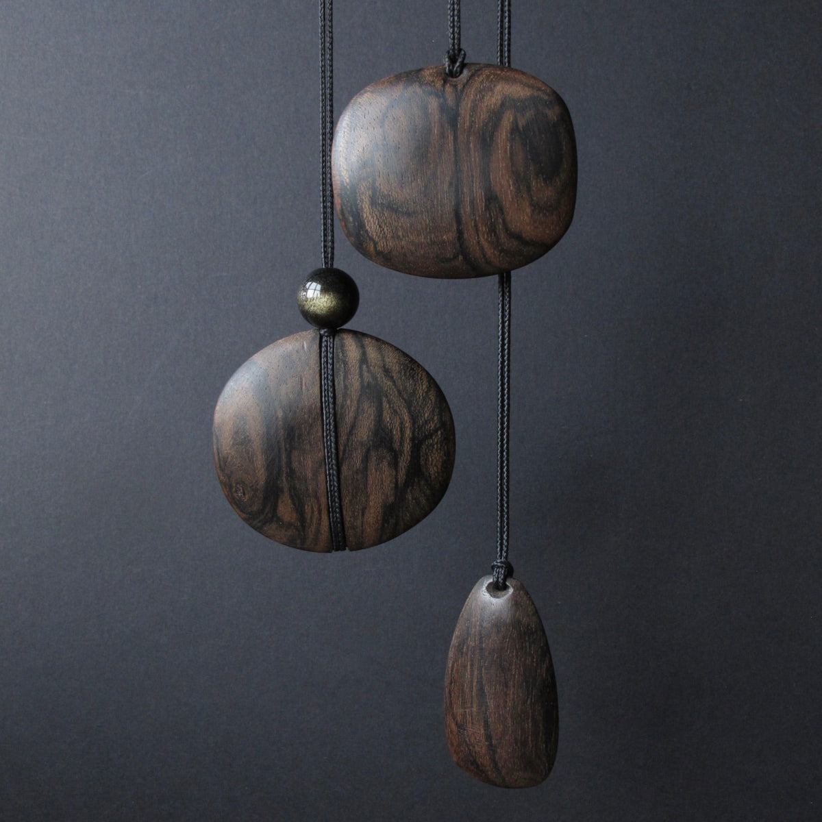 Three dark brown wooden hanging ornaments with natural wood grain patterns, suspended from black cords against a dark gray background.