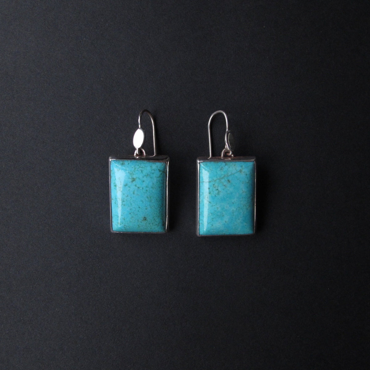 A pair of rectangular turquoise drop earrings with silver-toned metal borders and fishhook backings, displayed against a dark textured background.